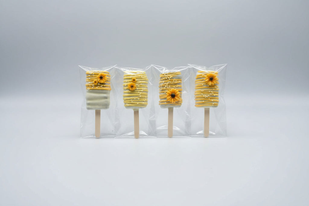 Four decorated cakesicle bars wrapped in clear cellophane sleeves, arranged vertically on a mottled gray and black stone surface. Each cakesicle has a white base with yellow drizzle, white chocolate stripes, and a small yellow flower decoration made by Honey, Listen!