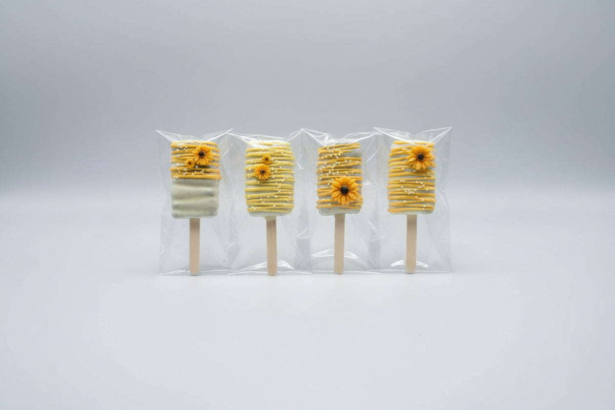 Four decorated cakesicle bars wrapped in clear cellophane sleeves, arranged vertically on a mottled gray and black stone surface. Each cakesicle has a white base with yellow drizzle, white chocolate stripes, and a small yellow flower decoration made by Honey, Listen!