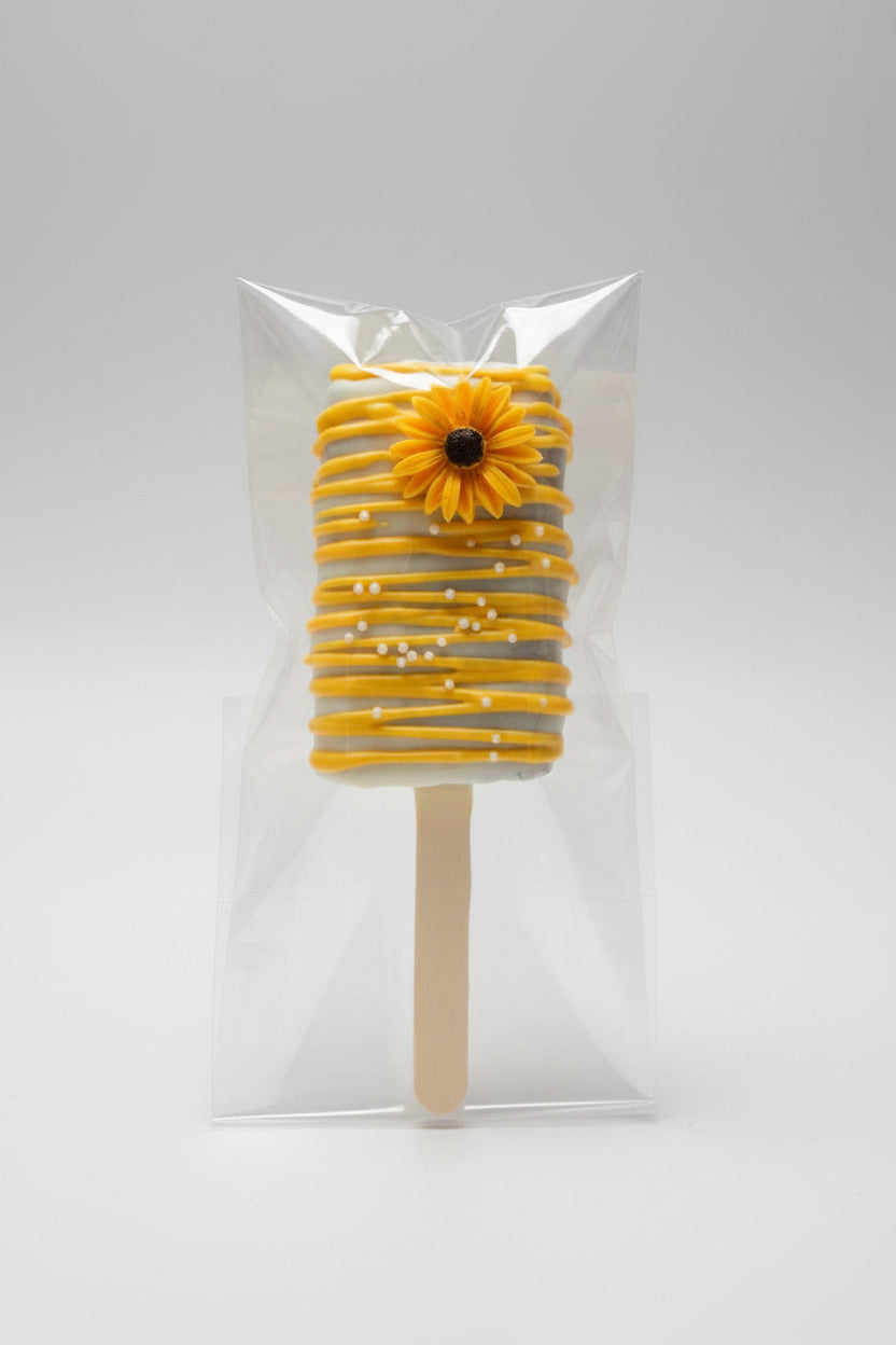 A yellow and white striped cakesicle with a small yellow flower decoration on top, made by Honey, Listen!, and sealed in a clear plastic bag