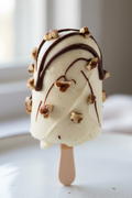 A close-up of a single cakesicle on a wooden stick, coated in creamy off-white chocolate with drizzles of dark chocolate and chopped nuts, set against a softly blurred light background.