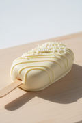 A single chocolate-covered cakesicle on a wooden board with a pale gray background. The cakesicle is coated in smooth white chocolate with decorative drizzle and sprinkles, attached to a wooden stick.