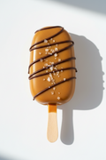 A caramel-topped cakesicle with chocolate drizzle and coarse salt crystals on a wooden stick, photographed on a clean white surface with a strong shadow.